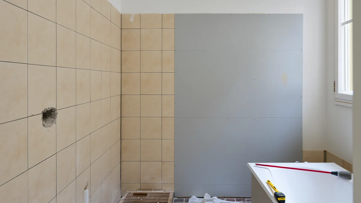Älteres gefliestes Bad im Altbau, eine Wand bereits mit neuer grauer Wandverkleidung überbaut, Wasserwaage und Maßband auf einer Arbeitsplatte