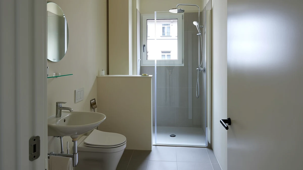Schmales kleines Bad mit Dusche hinter Klarglas am Fenster, Wand-WC vor einer halbhohen Vorwand mit Ablage und kleinem Waschtisch mit rundem Spiegel